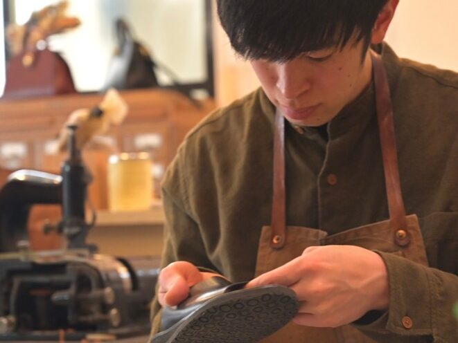 東京・荒川区の工房shiroで、職人が一針ずつ丁寧に革靴を仕立てる様子。初心者の方の初めてのオーダーを大切に、日本人の足に合う一生モノの履き心地を追求しています。エプロン姿の職人が黒い革を手に取り、静かな工房で作業に集中する風景です。