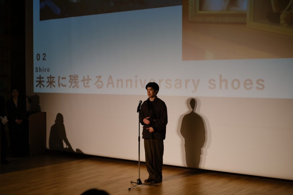 東京のイベント会場にて、初心者でも長く愛せる革靴について語る職人の姿。暗いステージでスポットライトを浴び、マイクを前に真剣な表情で「Anniversary shoes」への想いを伝える、プレゼンテーションの瞬間です。